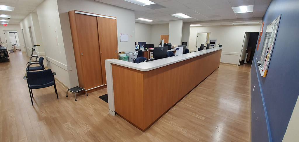 Long reception counter at a medical clinic, blue wall to the right, tall cabinets behind, long hallway to the left.